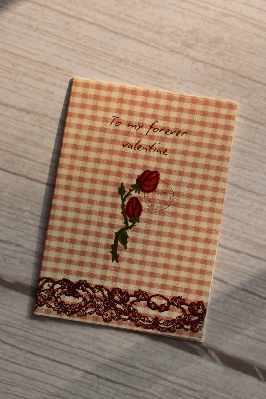 Forever Valentine Rose Greeting Card	Front cover of the Sunlit Hugs Forever Valentine Rose Greeting Card displayed in natural sunlight, featuring an orange gingham background, two vintage red roses, elegant calligraphy, and a printed dark lace border.