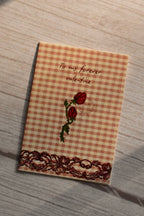 Forever Valentine Rose Greeting Card	Front cover of the Sunlit Hugs Forever Valentine Rose Greeting Card displayed in natural sunlight, featuring an orange gingham background, two vintage red roses, elegant calligraphy, and a printed dark lace border.