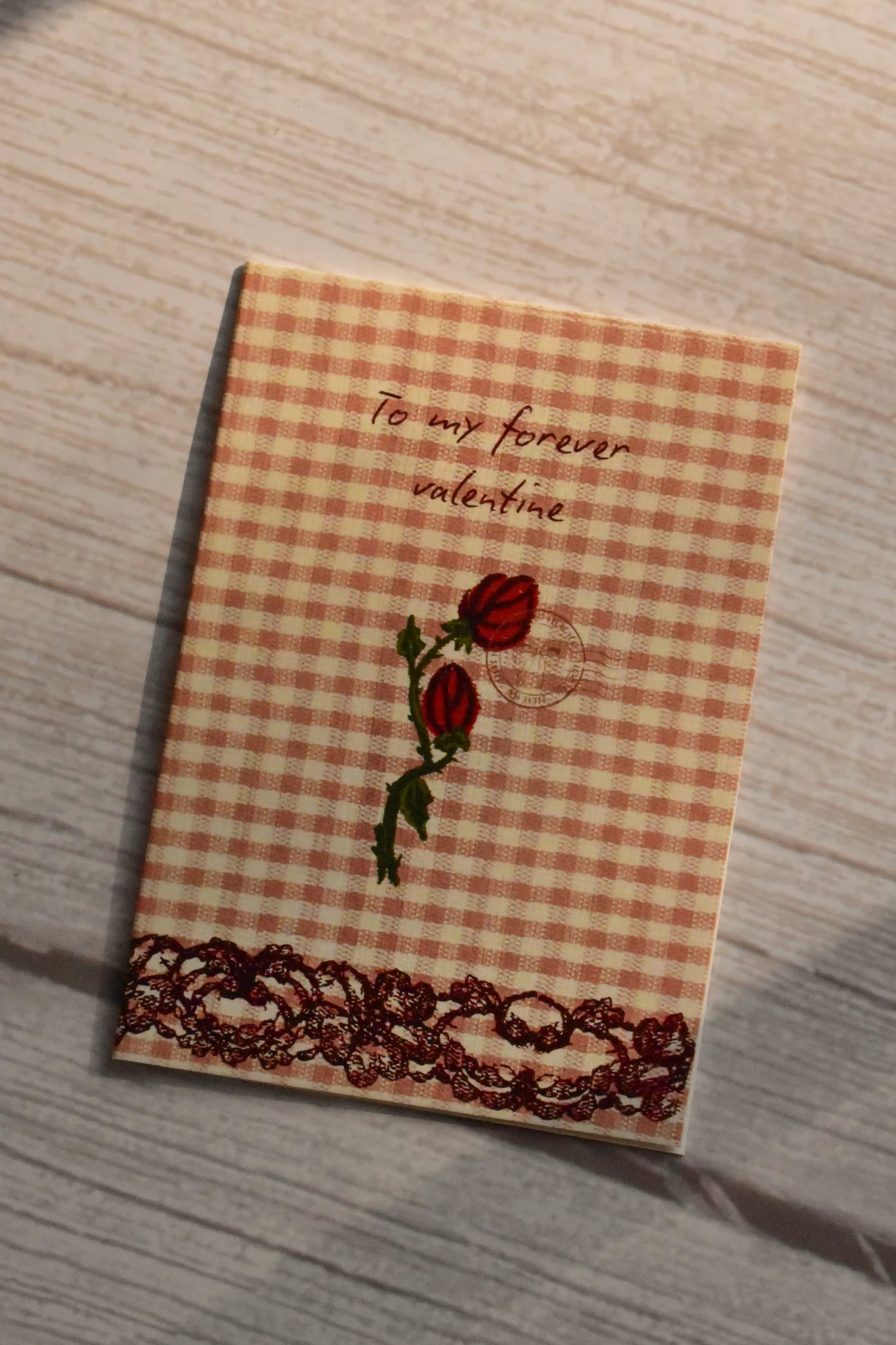 Forever Valentine Rose Greeting Card	Front cover of the Sunlit Hugs Forever Valentine Rose Greeting Card displayed in natural sunlight, featuring an orange gingham background, two vintage red roses, elegant calligraphy, and a printed dark lace border.