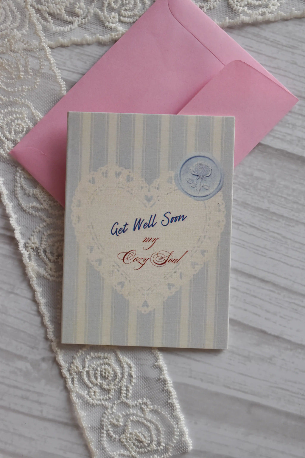 Front cover of the Sunlit Hugs Get Well Soon Cozy Soul Greeting Card resting on a pink envelope, featuring blue stripes, a heart doily design, and "Get Well Soon My Cozy Soul" text.