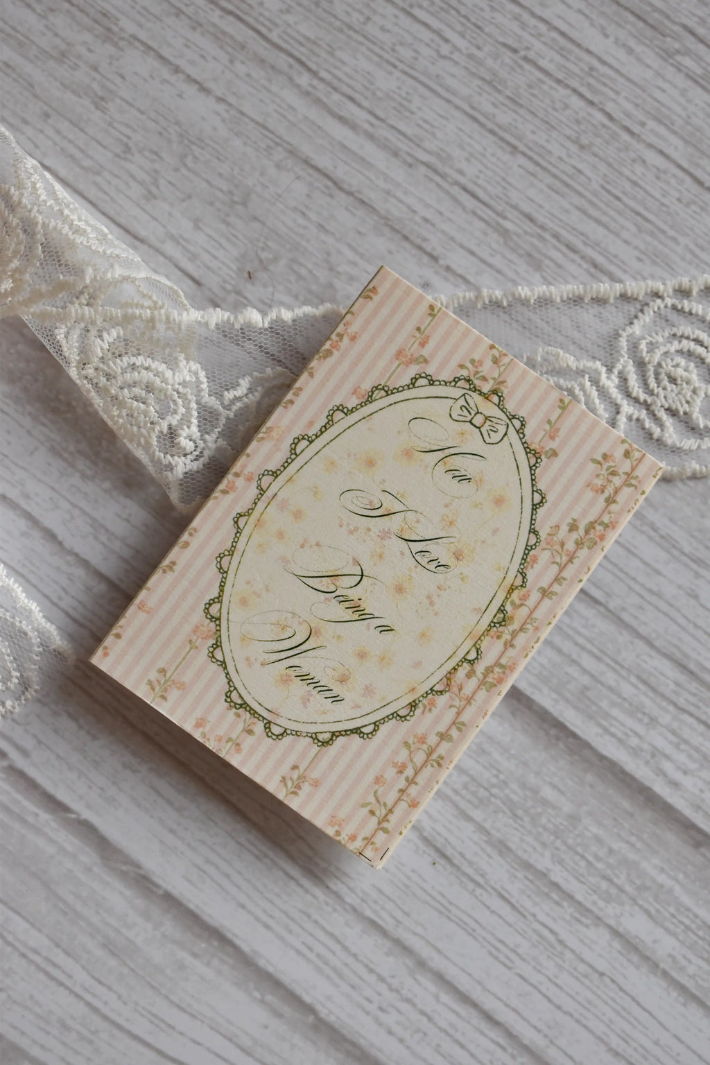 Front cover of the Sunlit Hugs How I Love Being a Woman Greeting Card featuring pink stripes, a vintage floral oval frame, and elegant typography next to white lace.