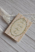 Front cover of the Sunlit Hugs How I Love Being a Woman Greeting Card featuring pink stripes, a vintage floral oval frame, and elegant typography next to white lace.