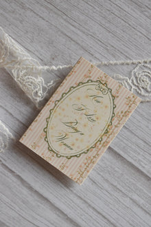 Front cover of the Sunlit Hugs How I Love Being a Woman Greeting Card featuring pink stripes, a vintage floral oval frame, and elegant typography next to white lace.