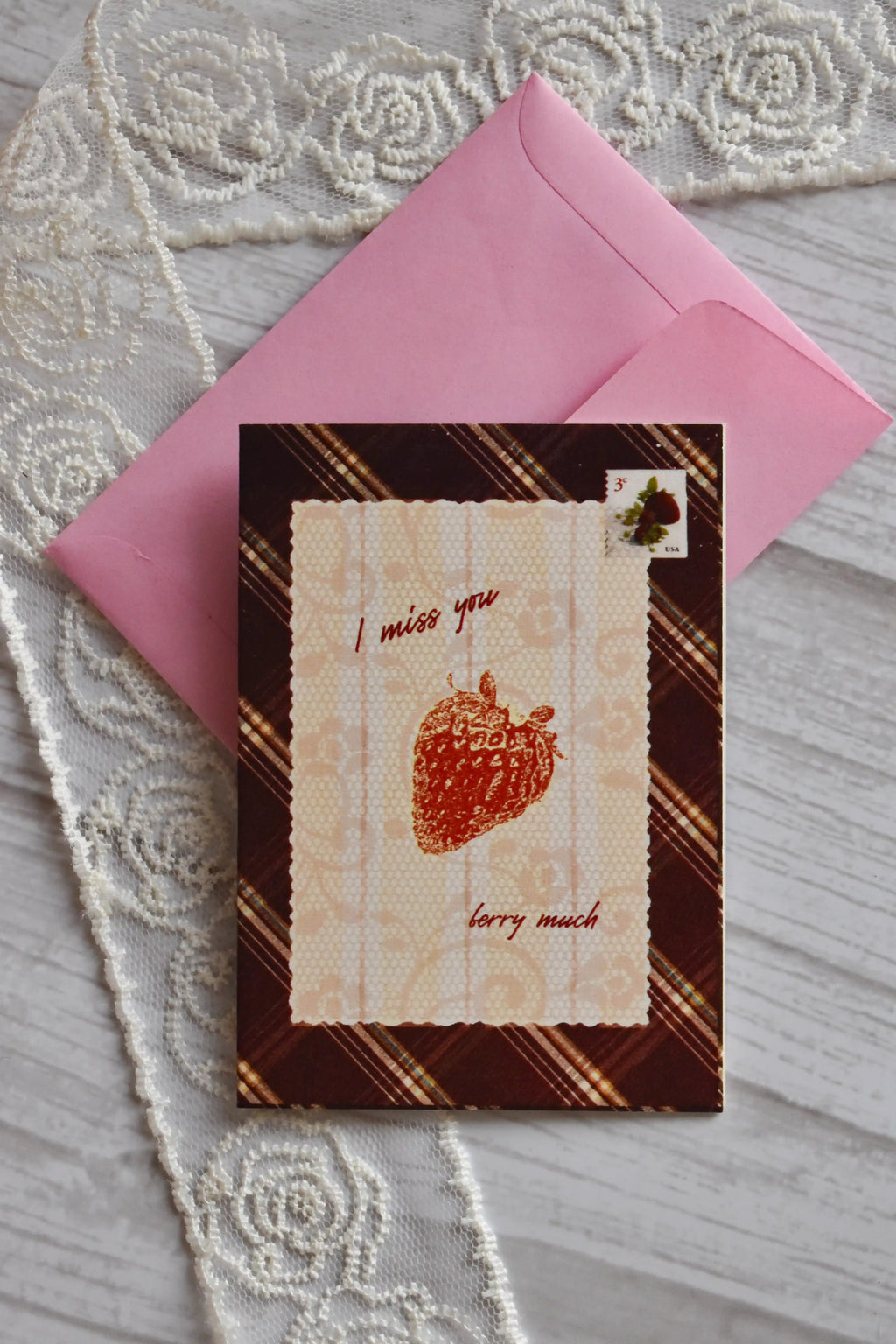 Front cover of the Sunlit Hugs I Miss You Berry Much Greeting Card resting on a pink envelope, featuring a brown plaid border, a strawberry graphic, and "I miss you berry much" text.