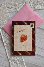 Front cover of the Sunlit Hugs I Miss You Berry Much Greeting Card resting on a pink envelope, featuring a brown plaid border, a strawberry graphic, and "I miss you berry much" text.