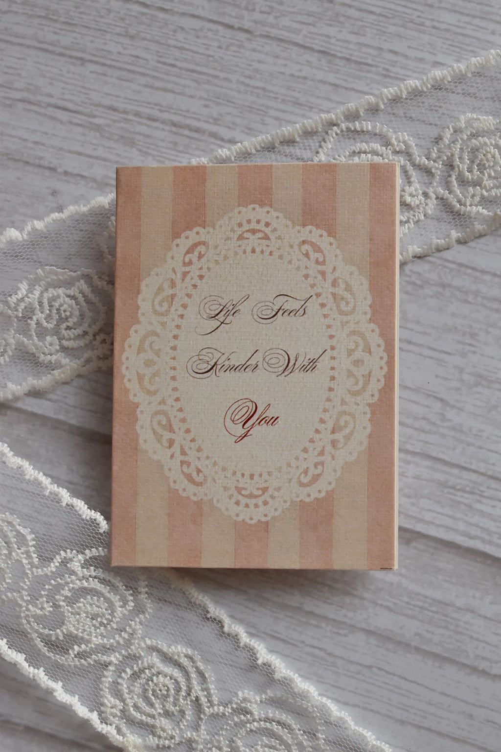 Front cover of the Sunlit Hugs Life Feels Kinder With You Greeting Card showing light pink stripes, a delicate white lace doily border, and romantic calligraphy.