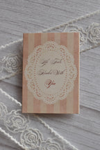 Front cover of the Sunlit Hugs Life Feels Kinder With You Greeting Card showing light pink stripes, a delicate white lace doily border, and romantic calligraphy.