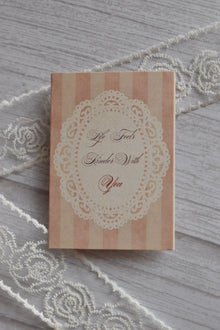 Front cover of the Sunlit Hugs Life Feels Kinder With You Greeting Card showing light pink stripes, a delicate white lace doily border, and romantic calligraphy.