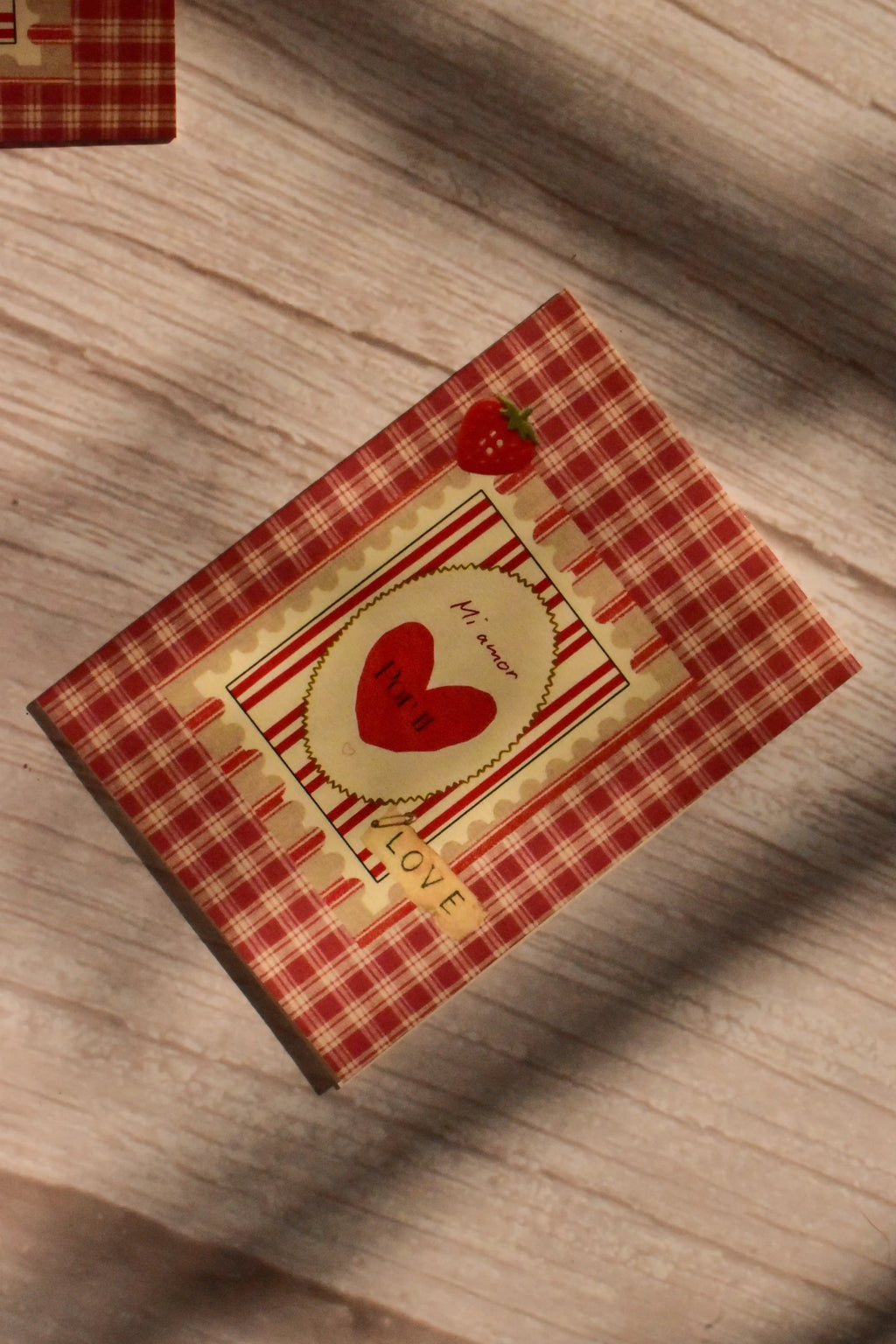 Flat lay front cover of the Sunlit Hugs Mi Amor Por Ti Greeting Card resting in sunlit shadows, featuring a vintage red plaid pattern, a heart illustration, a tiny strawberry, and a classic "LOVE" sticker detail.