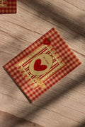 Flat lay front cover of the Sunlit Hugs Mi Amor Por Ti Greeting Card resting in sunlit shadows, featuring a vintage red plaid pattern, a heart illustration, a tiny strawberry, and a classic "LOVE" sticker detail.
