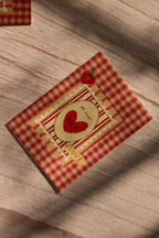 Flat lay front cover of the Sunlit Hugs Mi Amor Por Ti Greeting Card resting in sunlit shadows, featuring a vintage red plaid pattern, a heart illustration, a tiny strawberry, and a classic "LOVE" sticker detail.
