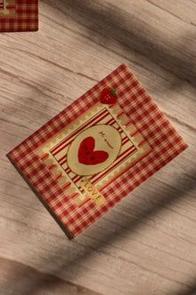 Flat lay front cover of the Sunlit Hugs Mi Amor Por Ti Greeting Card resting in sunlit shadows, featuring a vintage red plaid pattern, a heart illustration, a tiny strawberry, and a classic "LOVE" sticker detail.