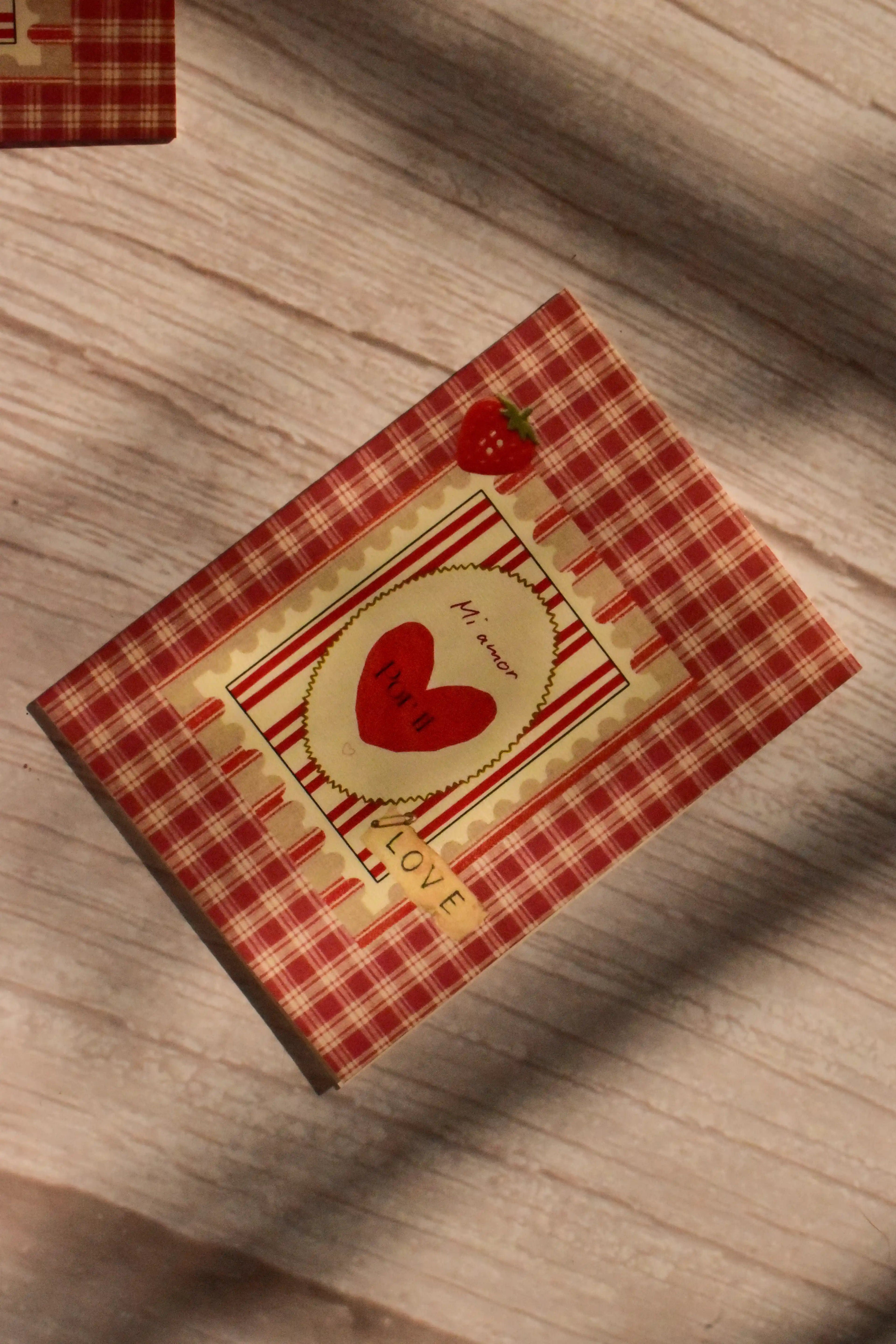 Flat lay front cover of the Sunlit Hugs Mi Amor Por Ti Greeting Card resting in sunlit shadows, featuring a vintage red plaid pattern, a heart illustration, a tiny strawberry, and a classic "LOVE" sticker detail.