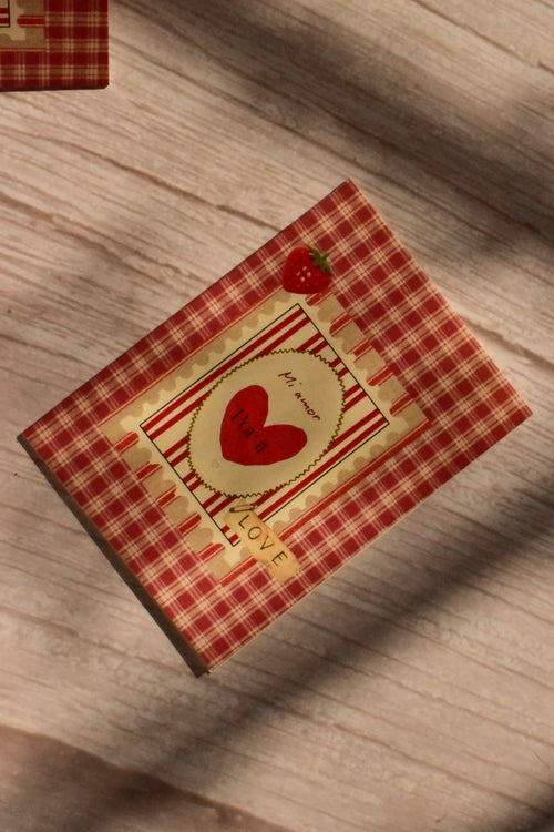 Flat lay front cover of the Sunlit Hugs Mi Amor Por Ti Greeting Card resting in sunlit shadows, featuring a vintage red plaid pattern, a heart illustration, a tiny strawberry, and a classic "LOVE" sticker detail.