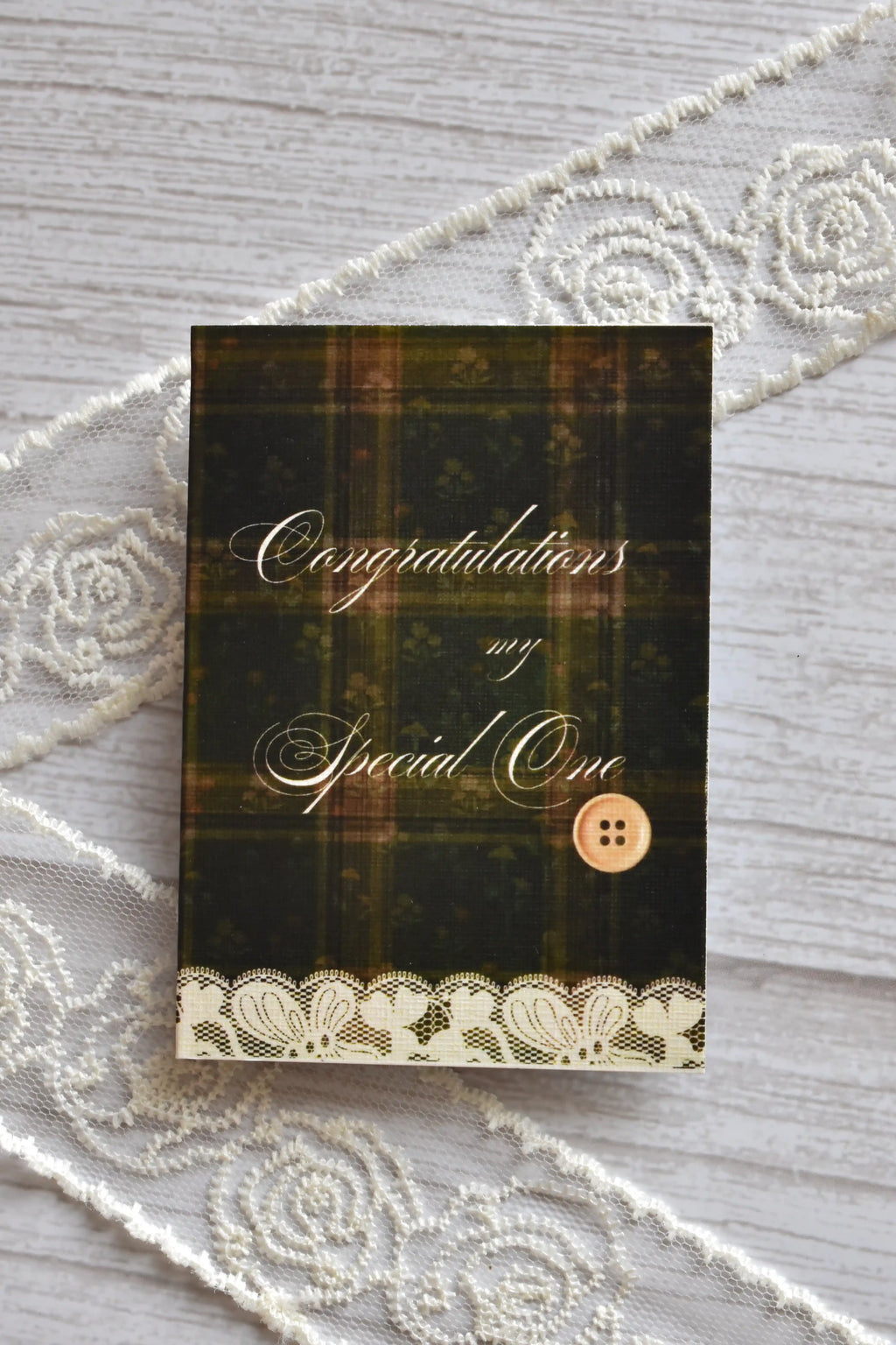 Front cover of the Sunlit Hugs Vintage Plaid Congratulations Greeting Card featuring dark green plaid, elegant calligraphy, a printed vintage button, and a white lace border.
