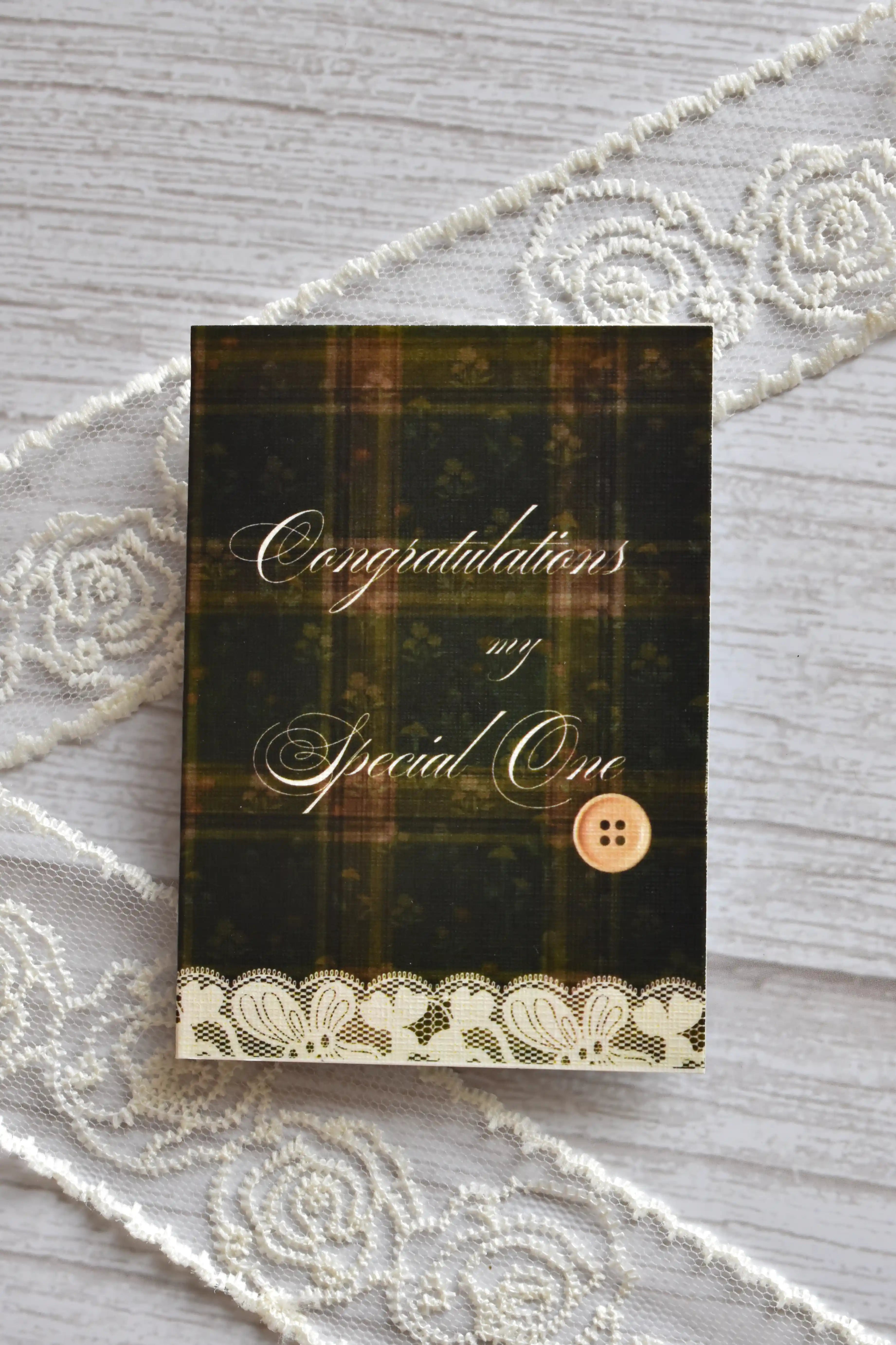 Front cover of the Sunlit Hugs Vintage Plaid Congratulations Greeting Card featuring dark green plaid, elegant calligraphy, a printed vintage button, and a white lace border.
