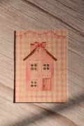 Front cover of the Sunlit Hugs Your Heart Is My Home Greeting Card featuring a charming cottagecore watercolor pink house, red ribbon bows, and romantic text on a wavy pink gingham background.