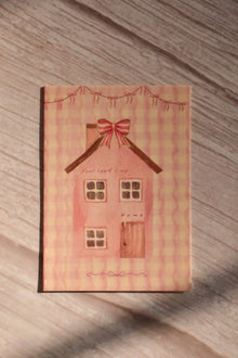Front cover of the Sunlit Hugs Your Heart Is My Home Greeting Card featuring a charming cottagecore watercolor pink house, red ribbon bows, and romantic text on a wavy pink gingham background.