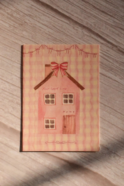 Front cover of the Sunlit Hugs Your Heart Is My Home Greeting Card featuring a charming cottagecore watercolor pink house, red ribbon bows, and romantic text on a wavy pink gingham background.