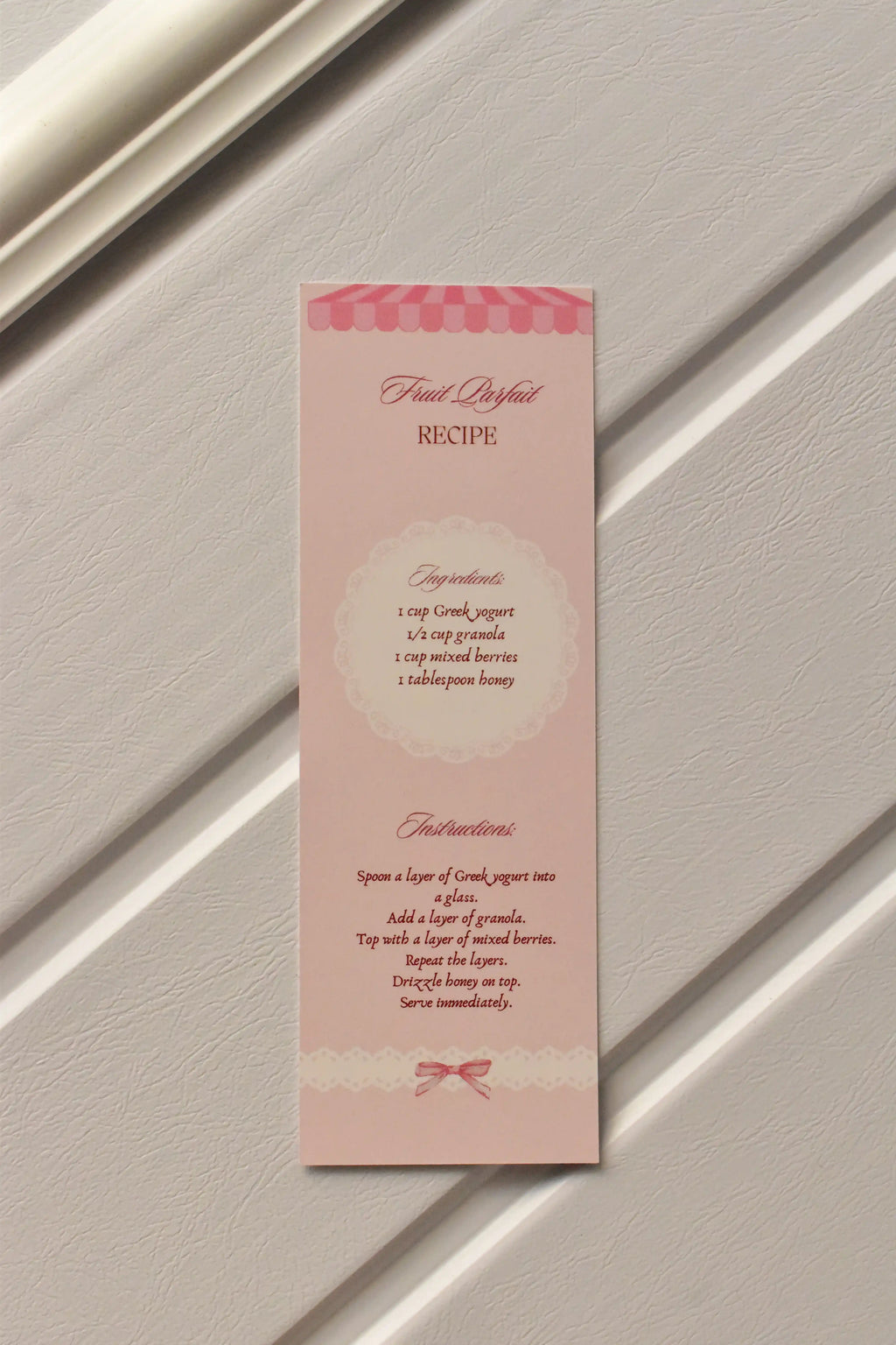 This image focuses on the practical element, displaying the detailed Fruit Parfait Recipe printed on the back of the pastel pink bookmark, ready for your kitchen inspiration.