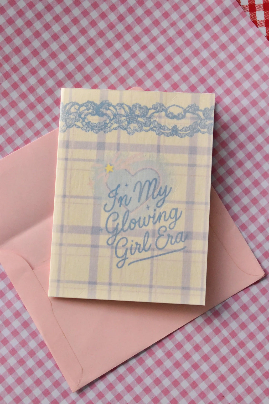 This image shows the front of the Glowing Girl Era greeting card. It features a light purple and white plaid design with a decorative blue lace border and the main blue script lettering message, sitting atop a pink envelope.