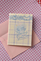 This image shows the front of the Glowing Girl Era greeting card. It features a light purple and white plaid design with a decorative blue lace border and the main blue script lettering message, sitting atop a pink envelope.