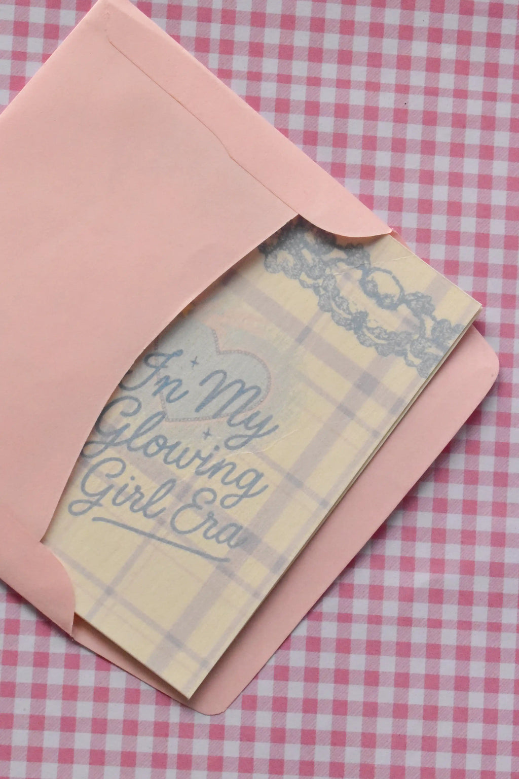This image demonstrates the card, with its light purple and white plaid design and blue text, being placed into the matching pink envelope. It highlights how the set arrives ready for sending a warm message.