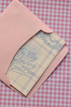 This image demonstrates the card, with its light purple and white plaid design and blue text, being placed into the matching pink envelope. It highlights how the set arrives ready for sending a warm message.