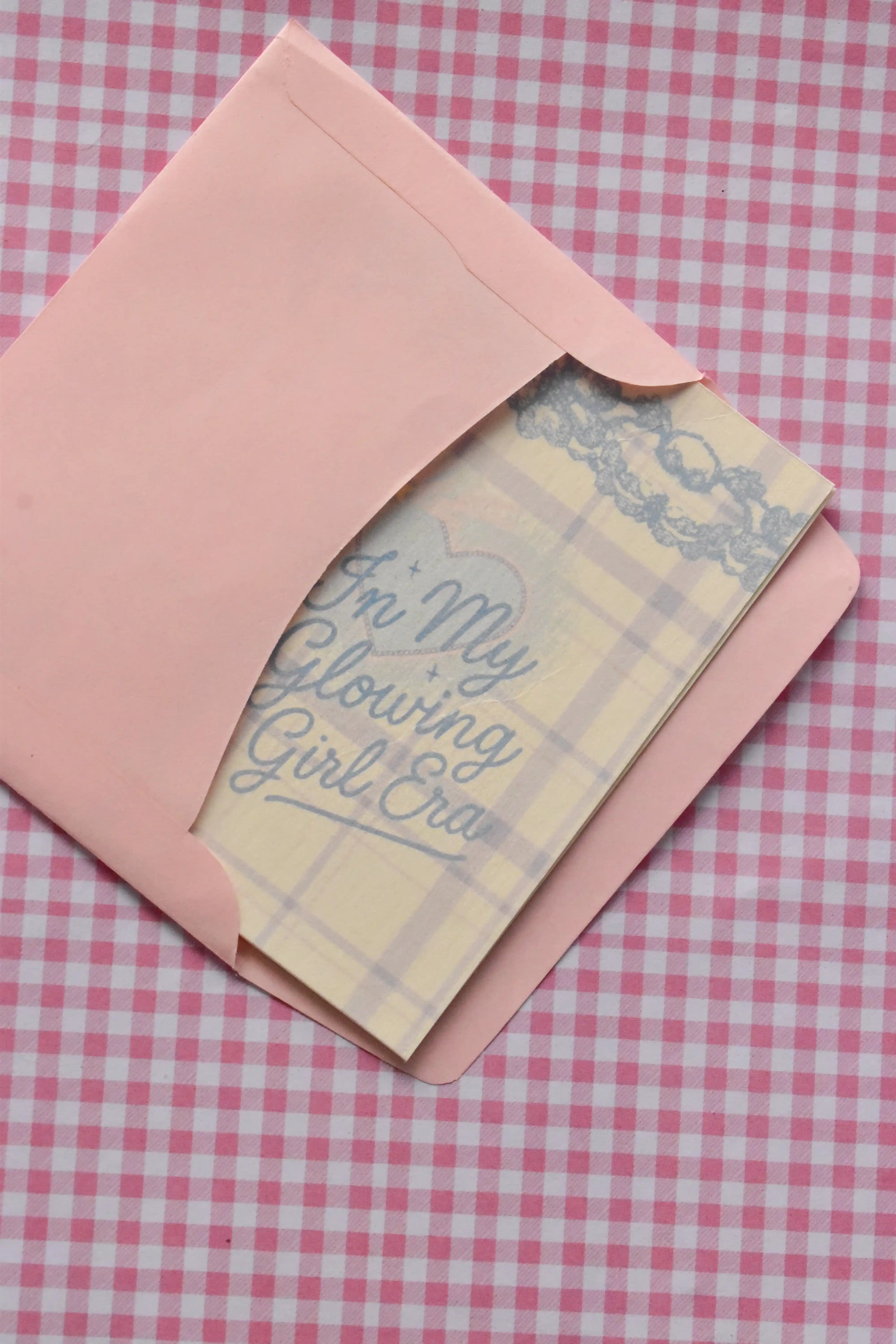 This view captures the Glowing Girl Era card being neatly tucked into the matching pink envelope. The light purple and white plaid card is partially visible, showing how the set arrives ready for sending a warm message.