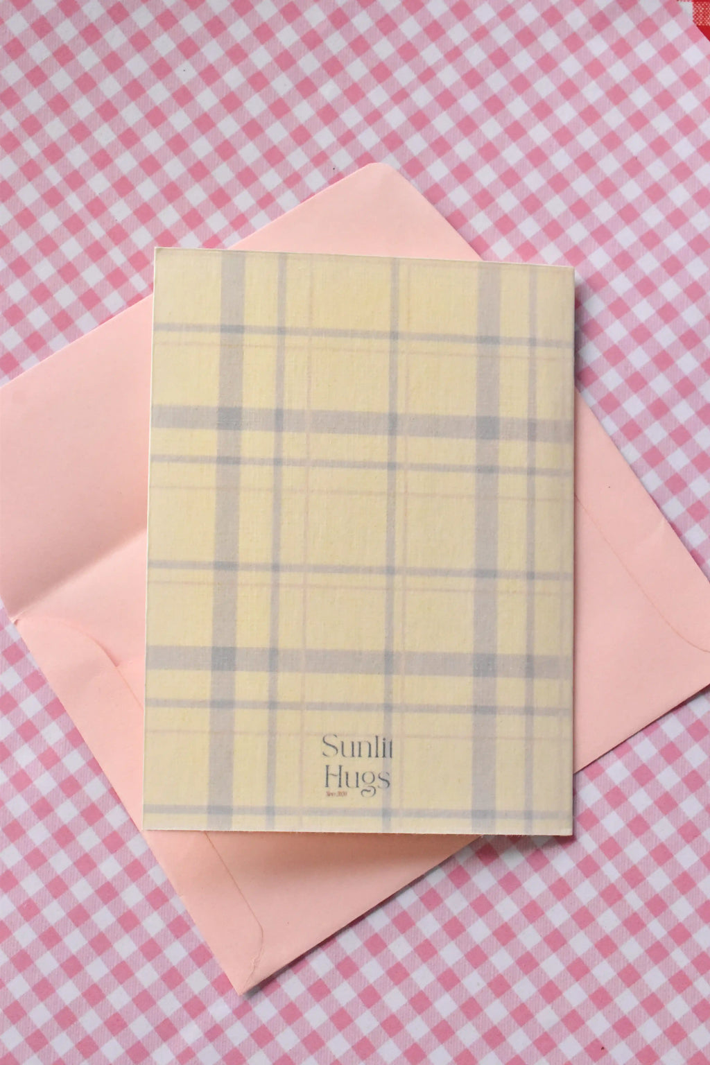 This image displays the back of the Glowing Girl Era card. The continuous light purple and white plaid pattern is visible, along with the Sunlit Hugs brand logo at the bottom, sitting inside a pink envelope.