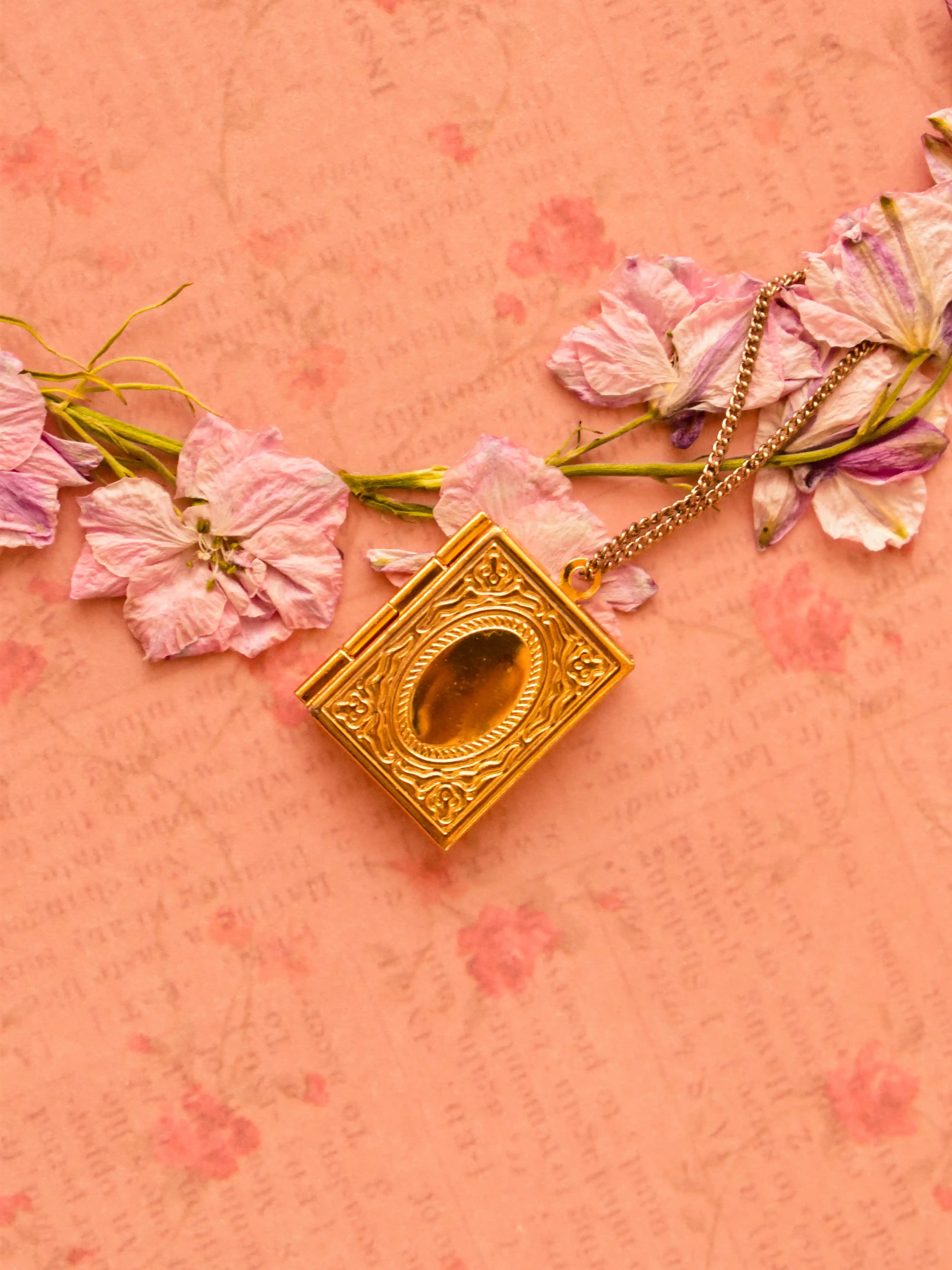 Detailed close-up of Sunlit Hugs’ golden book pendant, closed and placed on a romantic, handwritten-style pink paper.