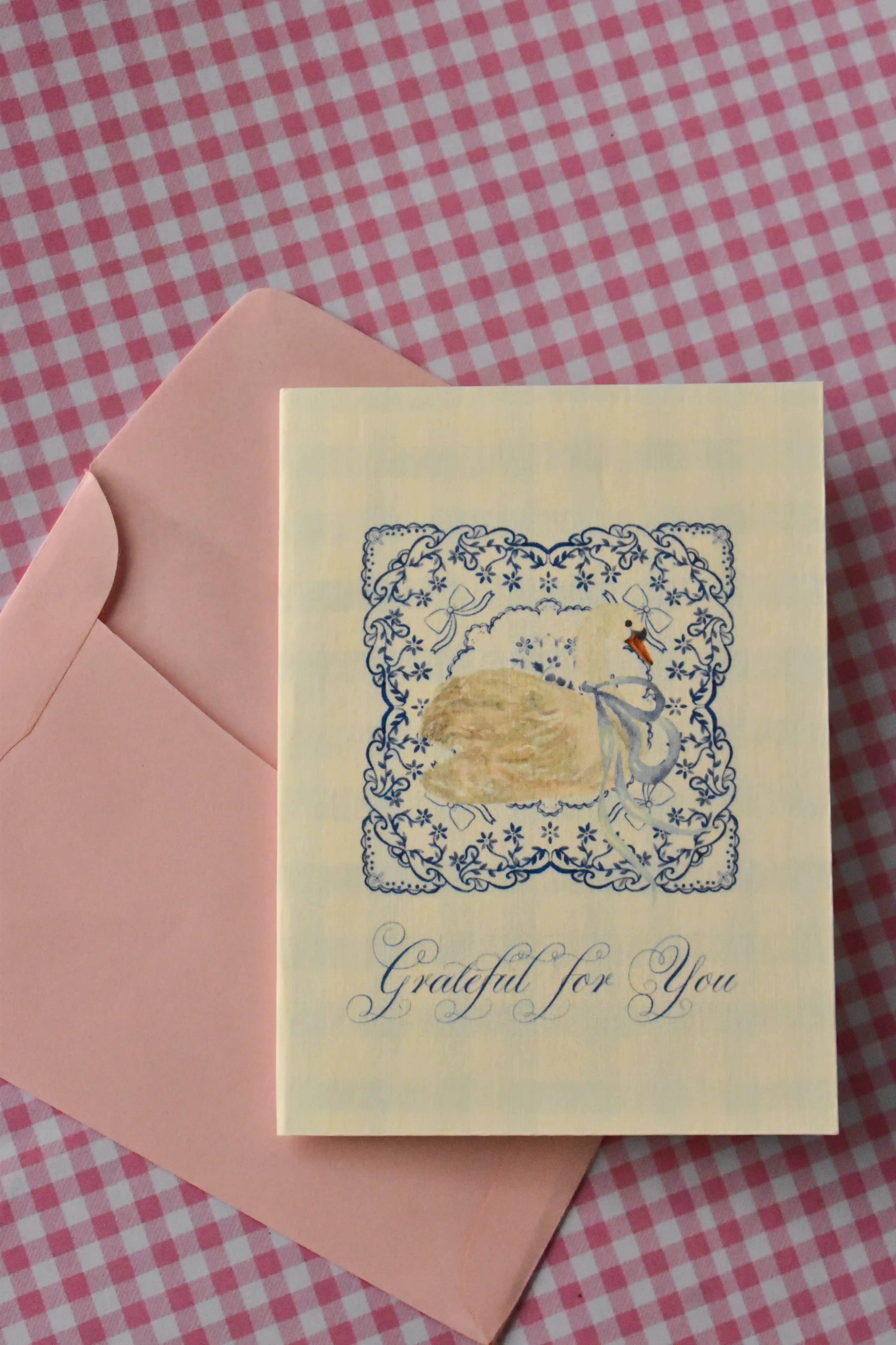This image shows the front of the Grateful For You card, capturing its delicate blue lace and swan design. The serene look sets a warm tone, ready for your note of appreciation, with the pink envelope nearby.