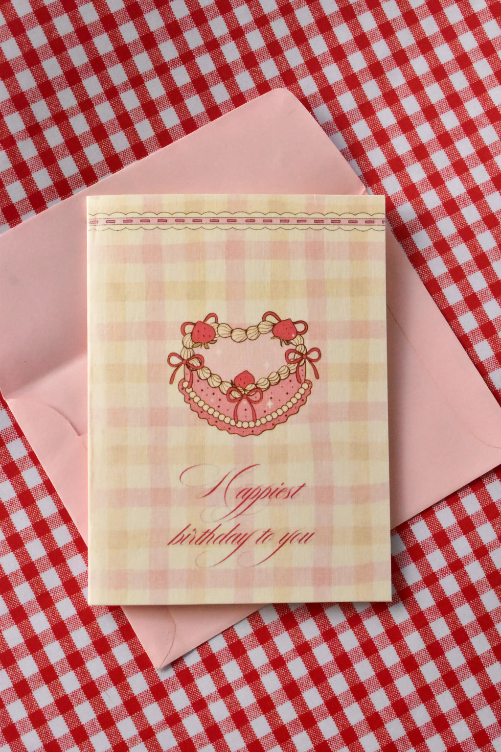 This image showcases the front of the Happiest Birthday to You card, highlighting the sweet pink heart-shaped cake design and the cosy pink and cream gingham pattern, ready to send warm wishes.