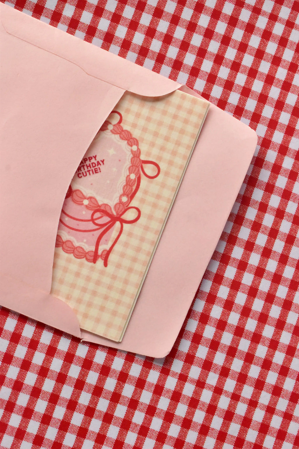 This view captures the Happy Birthday Cutie card being gently placed into the pink envelope. It demonstrates the presentation, with the sweet pink heart-shaped cake illustration peeking out as it awaits its recipient.