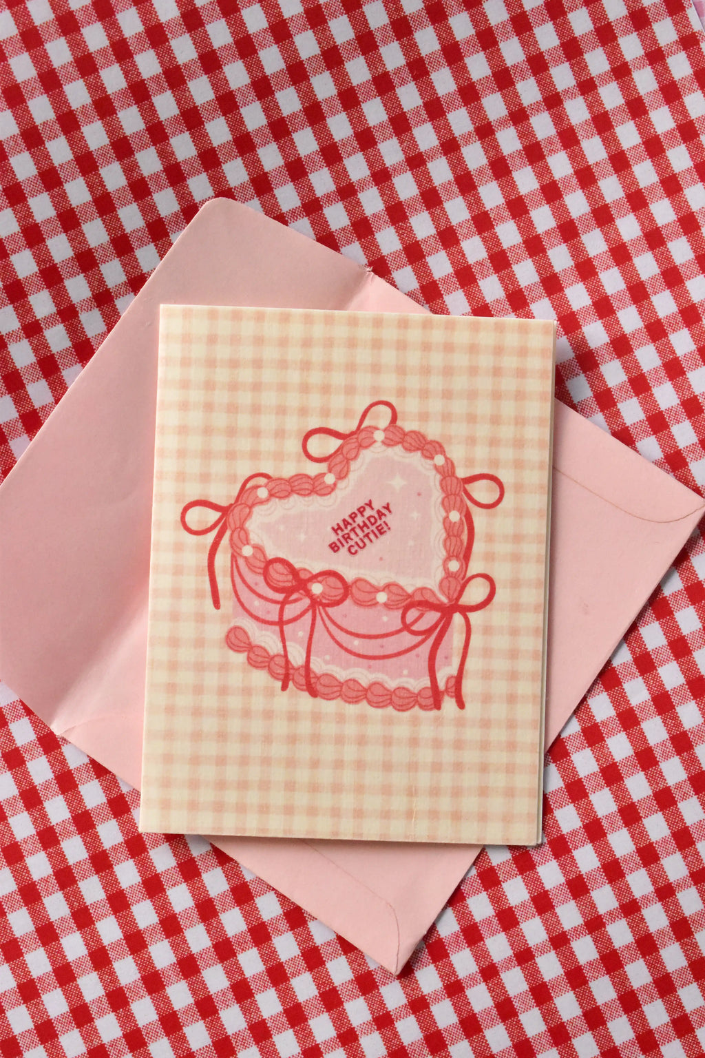 A close-up view highlighting the sweet pink heart-shaped cake design and the cosy pink gingham pattern of the 'Happy Birthday Cutie' card from the set.