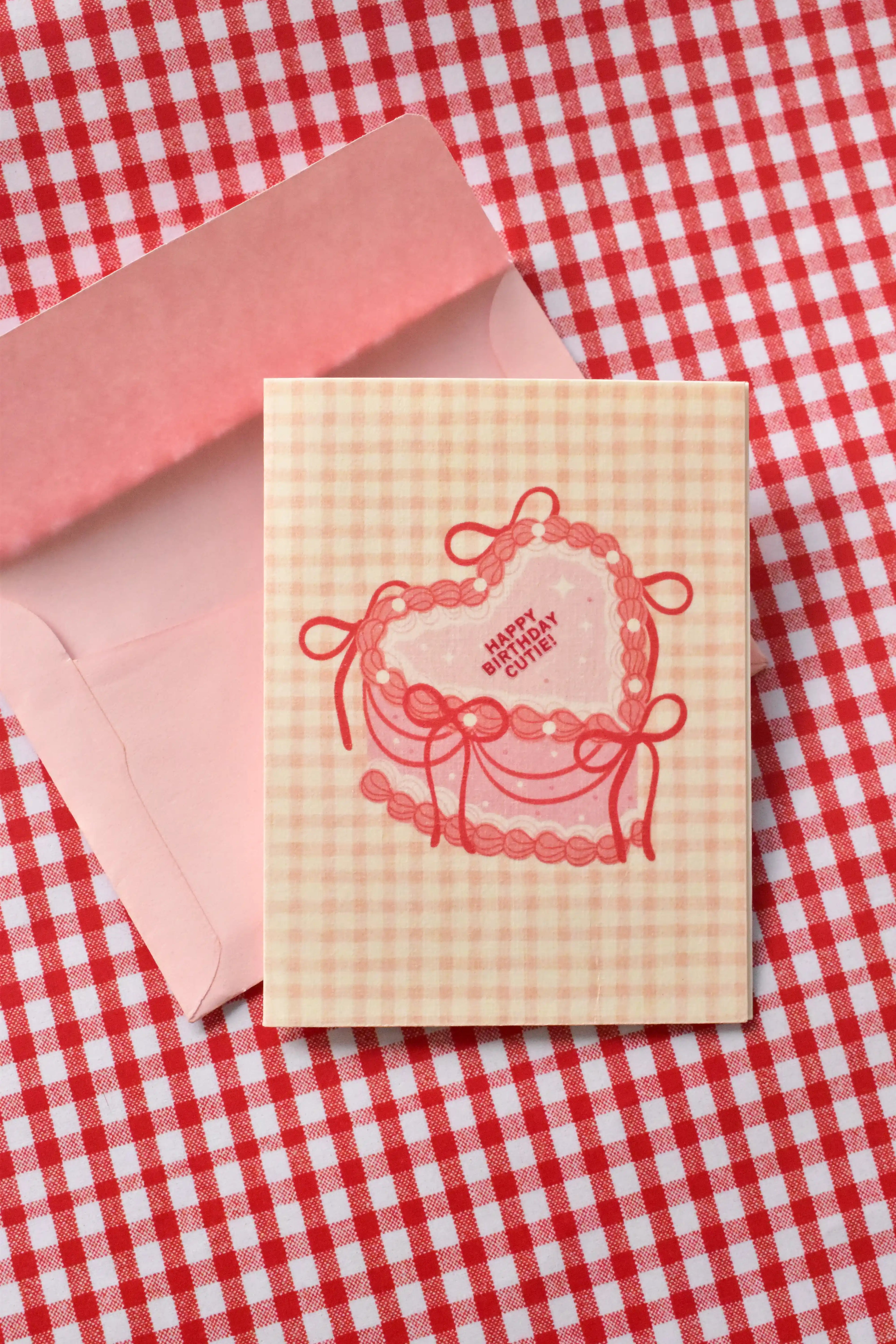 This image showcases the front of the Happy Birthday Cutie card, highlighting the sweet pink heart-shaped cake design and the cosy pink and cream gingham pattern, ready to send warm wishes.
