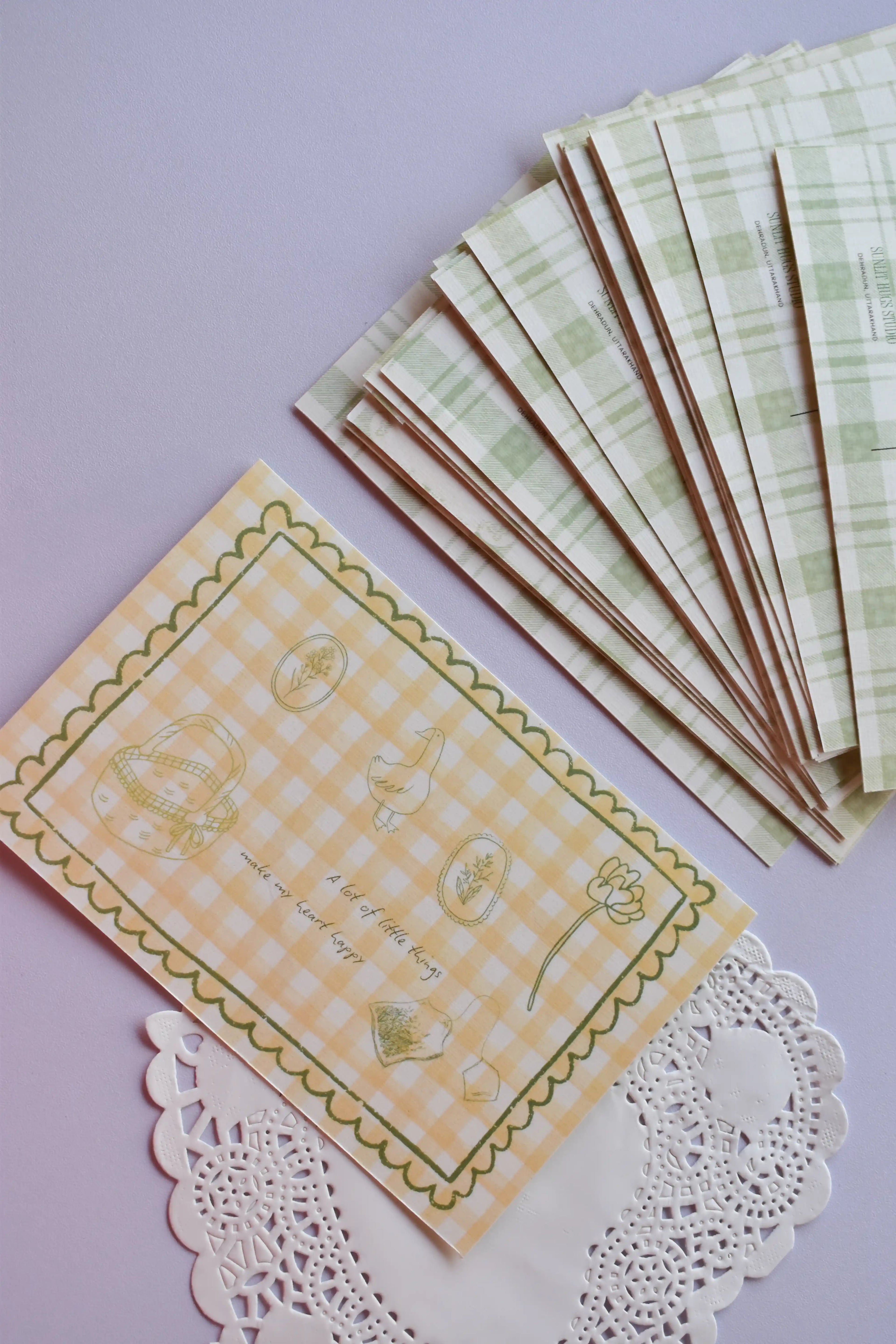A clear view of the 'A lot of little things make my heart happy' postcard front, featuring yellow wide gingham and line-art sketches. A stack of the green plaid reverse sides, showing the duck and address lines, is arranged neatly above it.