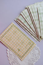 A clear view of the 'A lot of little things make my heart happy' postcard front, featuring yellow wide gingham and line-art sketches. A stack of the green plaid reverse sides, showing the duck and address lines, is arranged neatly above it.