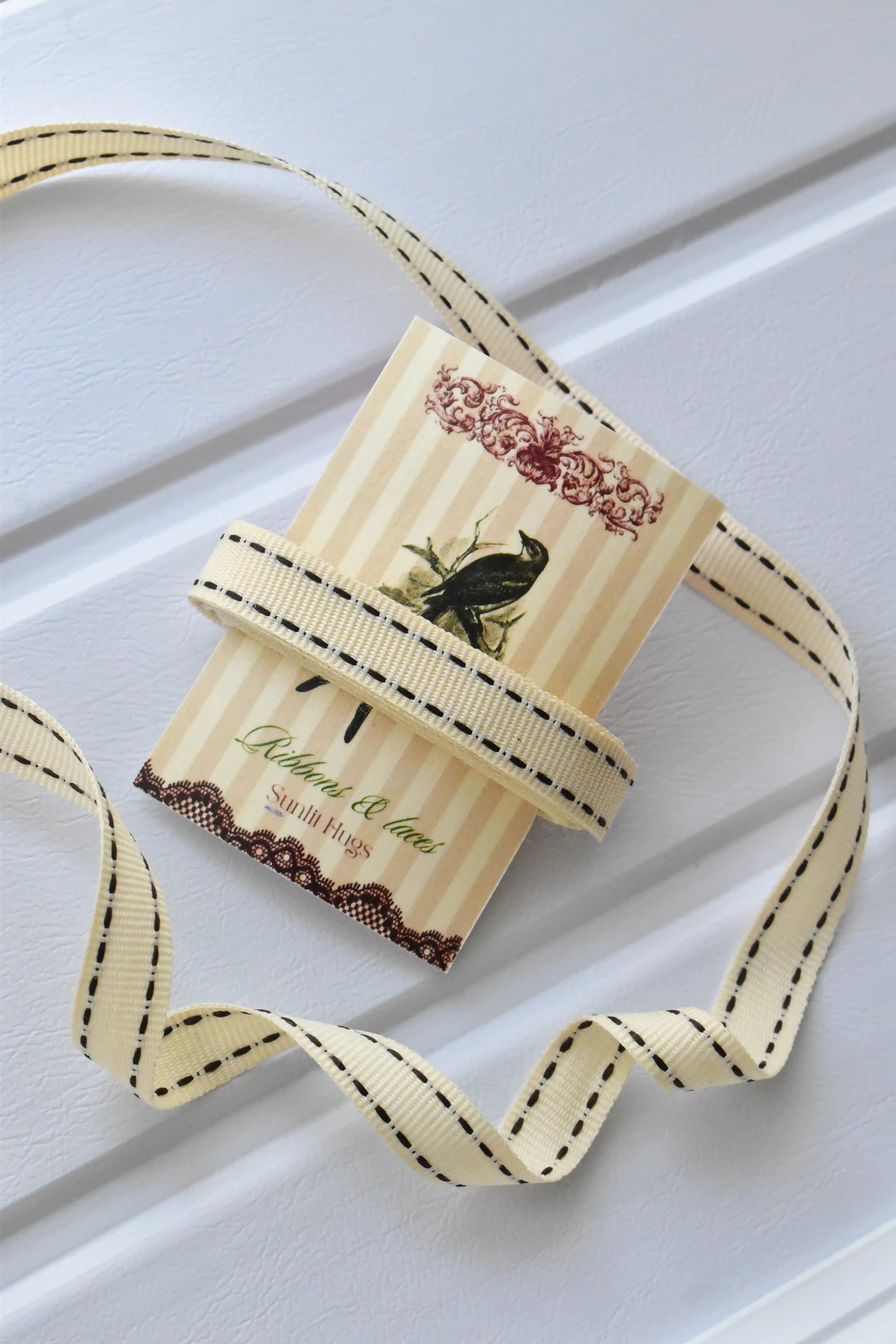 This image displays the Ivory Stitch ribbon, showcasing its cream colour and delicate brown dashed stitching. A long strand is loosely unravelled to highlight the full design, with a section wrapped around a "Sunlit Hugs" brand tag.