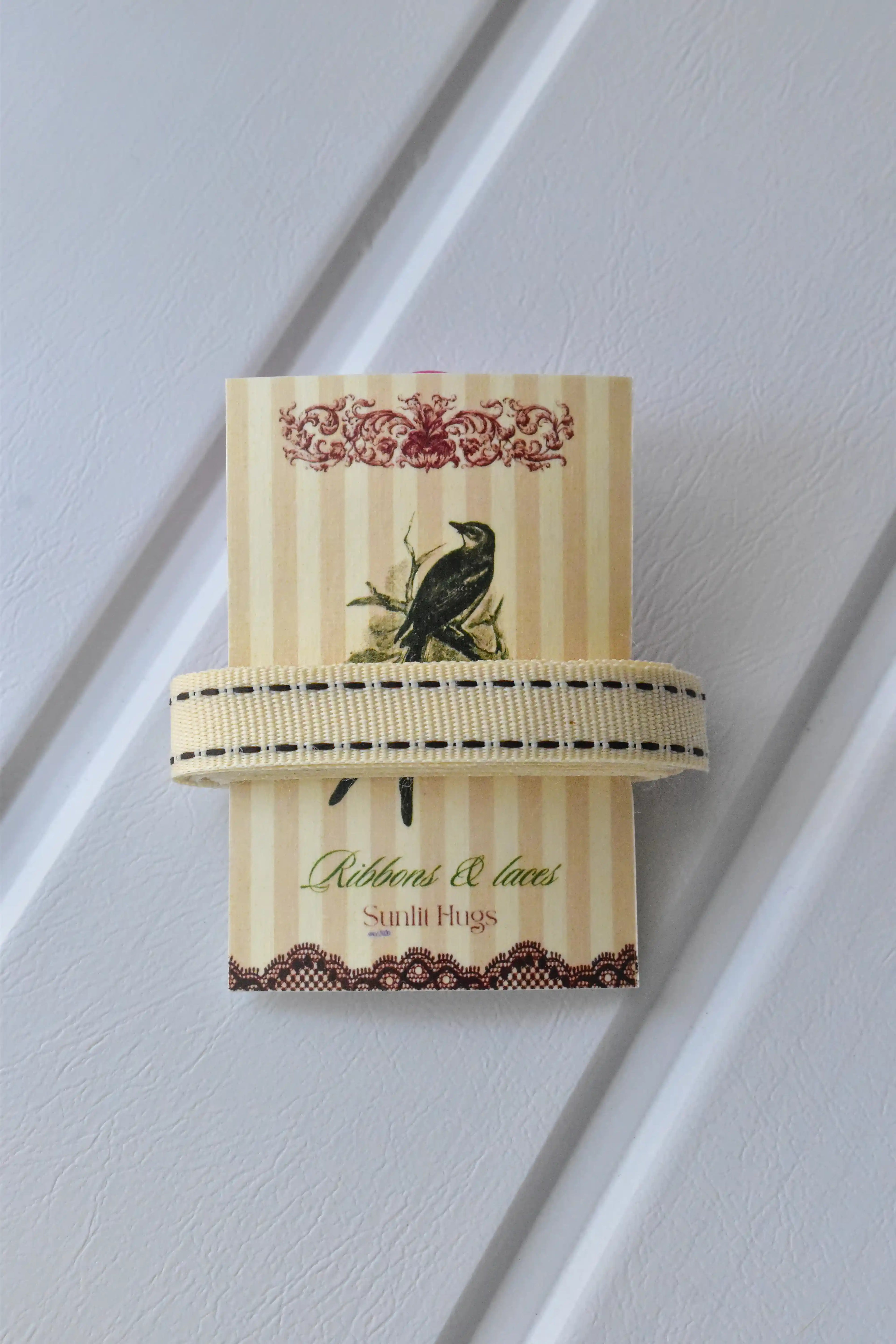 This detailed, head-on view of the Ivory Stitch ribbon highlights its soft cream colour and the contrasting brown dashed stitching. The ribbon is wrapped tidily around the "Sunlit Hugs" brand tag to show its precise design.