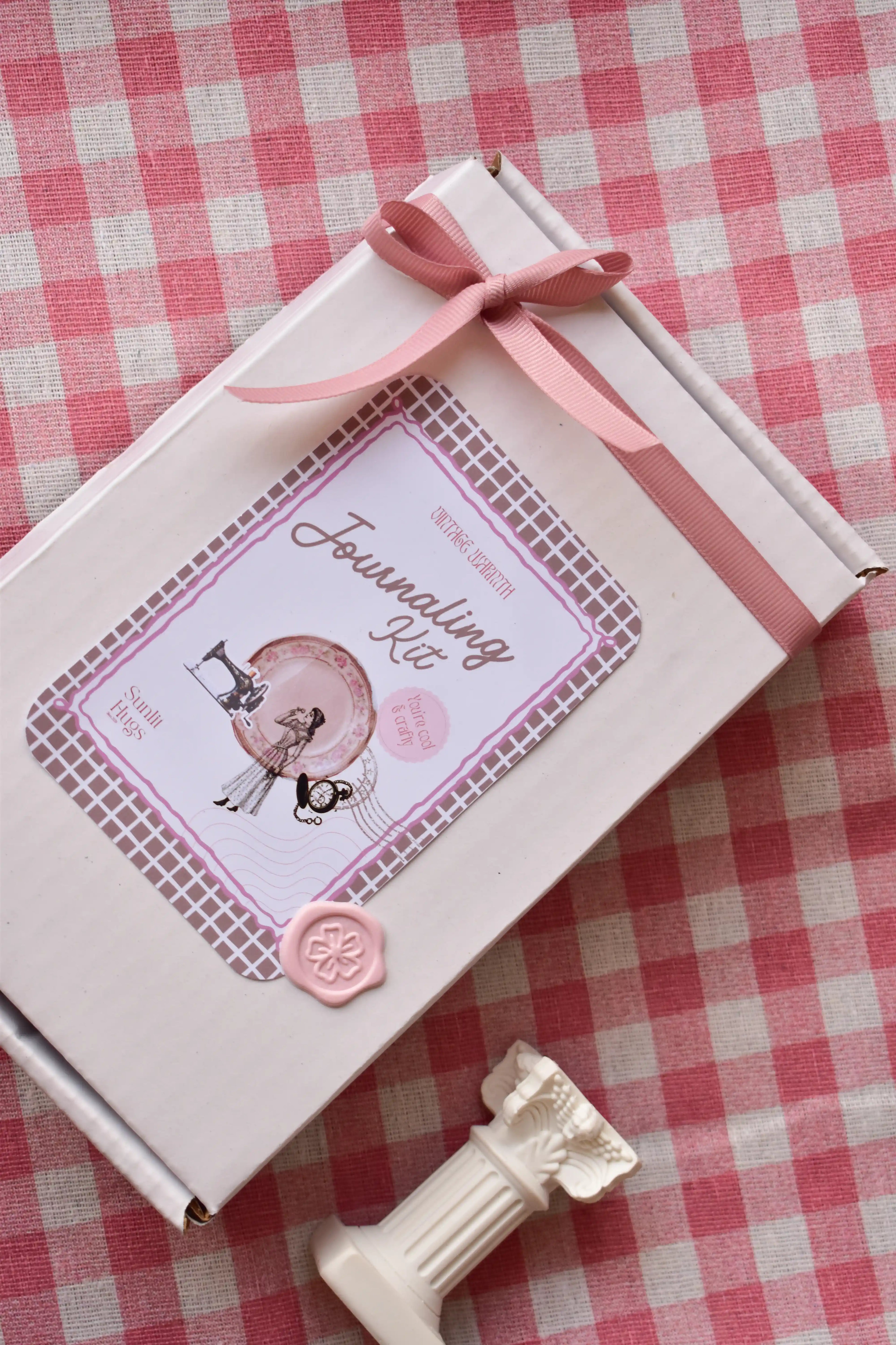White Journaling Kit box tied with a pink ribbon, sealed with a pink wax seal, resting on a pink and white checkered background.