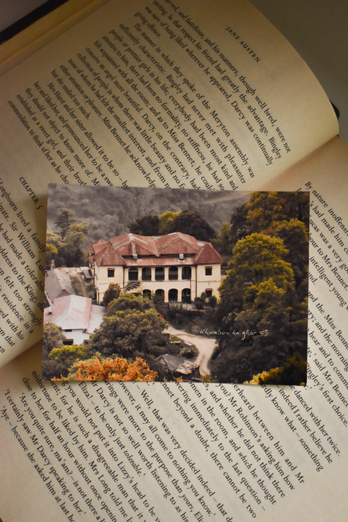 Flat lay of the 3x4 inch 'Khwabon Ka Ghar' Barlowganj Photo Print tucked inside a classic novel, featuring fine art mountain photography of a vintage estate nestled among lush green and autumn-tinged trees.