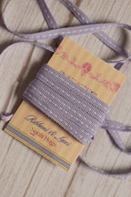 Lilac Stitches Narrow Ribbon shown with loose ribbon strands around the product card, highlighting its soft purple tone and white stitched detail for crafts and packaging.