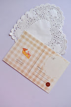 Close-up of the reverse side of a postcard on a white heart-shaped paper doily. The postcard has a tan and cream wide gingham pattern and features a small illustration of a slice of pie. It has sections for the address, postage, and a decorative brown button.