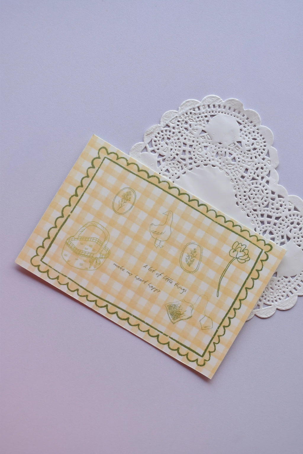 A horizontal postcard featuring a yellow and white wide gingham pattern with whimsical line-art sketches (a basket, a duck, and flowers) and the quote, "A lot of little things make my heart happy." A stack of the green gingham reverse side of the postcards is visible behind it.