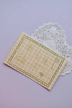 A horizontal postcard featuring a yellow and white wide gingham pattern with whimsical line-art sketches (a basket, a duck, and flowers) and the quote, "A lot of little things make my heart happy." A stack of the green gingham reverse side of the postcards is visible behind it.