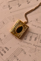A close-up of the closed golden alloy locket, showcasing the intricate, book-style engraved design on the front, set against warm, nostalgic sheet music. (Note: Chain is for visual purposes only).