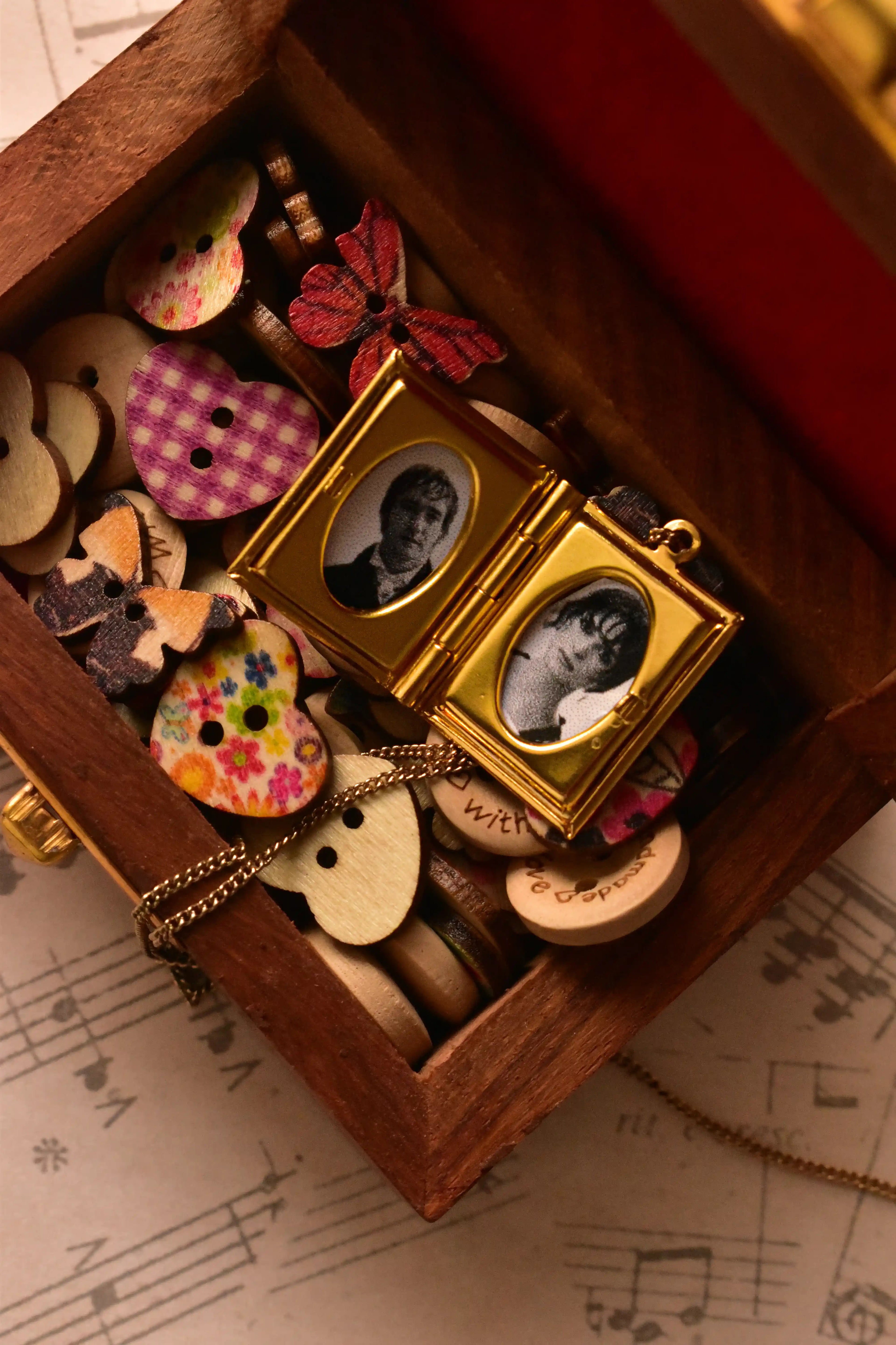 A warm display shot showing the open golden alloy locket surrounded by whimsical, colourful buttons and wooden charms, illustrating its personal, treasured value.