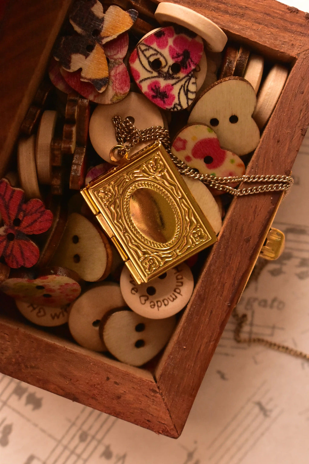  A warm, styled shot of the closed golden alloy locket tucked inside a wooden box surrounded by nostalgic wooden and patterned buttons, highlighting its appeal as a romantic, vintage treasure.