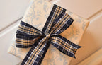 This image displays the Midnight Plaid ribbon, showcasing its rich navy and white plaid design and woven texture. A long strand is loosely unravelled to highlight the full pattern, with a section wrapped around a "Sunlit Hugs" brand tag.